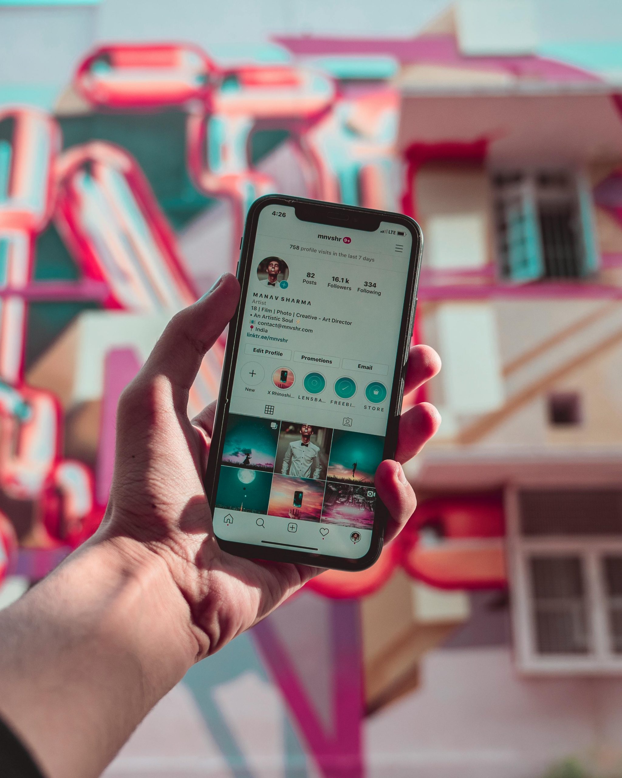 A hand holds a smartphone displaying social media, set against a colorful urban mural.